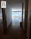 Empty hallway with wooden floors leading to a bright room with large windows. Debris scattered on the floor.
