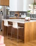 Modern kitchen with wood island, white bar stools, stainless steel fridge, geometric backsplash, and hanging plants.