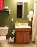 Green bathroom with wooden vanity, mirror, white sink, toilet, and towel rack. Basket with toilet paper on floor.