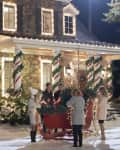 People decorating a red sleigh with greenery and lights in front of a festive house with candy cane columns.