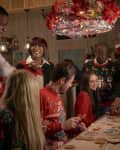 Group of people in festive sweaters decorating Christmas cookies under a chandelier with red poinsettias.