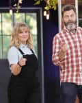 Two people standing indoors, one in a plaid shirt and the other in overalls, both pointing at the camera.