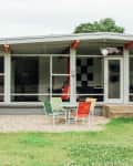 Mid-century modern house exterior with large windows, orange beams, and colorful patio chairs on gravel.