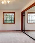 Empty room with beige carpet, two windows, ceiling fan, and mirrored closet doors.