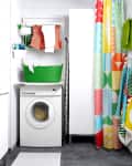 Laundry room with washing machine, colorful shower curtain, green laundry basket, and clothes hanging to dry.