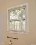 Bathroom with white tiled shower, window with blinds, and a metal grab bar above a toilet paper holder.