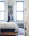 Bedroom with white bedding, colorful rug, and clothes hanging under a window with shutters.