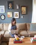 Woman using a laptop on a beige sofa, surrounded by eclectic wall art, with a wooden coffee table holding books and a candle.