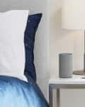 Bedside table with a lamp, smart speaker, succulent, and watch next to a bed with blue and white bedding.