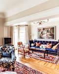 Elegant living room with patterned armchairs, blue velvet sofa, red and blue rugs, and large windows with beige curtains.