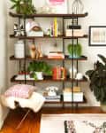 Bookshelf with plants, books, decor items, and a black lamp. A stool with a red patterned pillow and a white phone below.