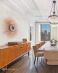 Elegant dining room with a long wooden table, beige chairs, chandelier, and large windows overlooking a cityscape.