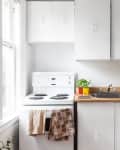 Small kitchen with white cabinets, electric stove, wooden countertops, potted plant, and a microwave with bananas on top.