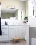 White bathroom with large mirror, three-light fixture, white cabinets, and a small wooden stool on tiled floor.