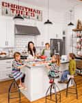 Family decorating gingerbread cookies in a white kitchen with a "Christmas Trees for Sale" sign above.