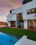 Modern white house with large windows, pool, and green lawn at sunset.