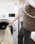 Woman loading patterned rug into front-load washing machine, holding a woven laundry basket.