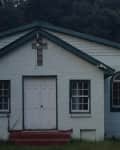 Small white brick church with green trim, arched windows, and a cross above the door.
