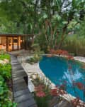 Mid-century modern house with large windows, lush garden, and a kidney-shaped pool surrounded by trees and plants.