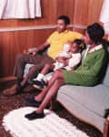 Family watching TV in a wood-paneled living room, sitting on a green sofa, child holding a doll.