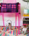Pink acrylic chair with a colorful cushion, next to a stack of books, a watering can, and a small table with plants.