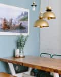 Dining area with a wooden table, gold pendant lights, framed beach photo, and eucalyptus in a vase.