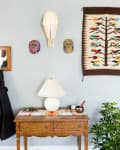 Wooden console table with lamp, plant, and decor under a mirror and colorful tapestry on a light blue wall.