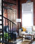 Loft-style living room with exposed brick, cream sofa, blue and yellow pillows, spiral staircase, and modern decor.
