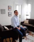 Man sitting on a brown sofa in a living room with framed art, cowhide rug, and a floor lamp.