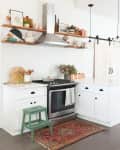 White kitchen with hex tile backsplash, open shelves, green step stool, copper mugs, and a colorful rug.
