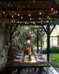 Outdoor dining area with wooden table, string lights, and floral centerpiece under a pergola surrounded by greenery.