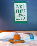 Green wall hanging with "Time Cools All Jets" above a bed with white pillows and an orange hat labeled "Boon."