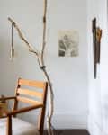 Wooden chair beside a branch lamp with hanging bulb, wall art, and wooden utensils on a white wall.