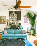 Eclectic living room with teal sofa, colorful pillows, large plants, brick fireplace, and vibrant artwork on walls.