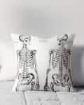 Skeleton print pillow on a bed with a gray quilt, next to a small table with a potted plant.