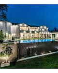 Modern two-story house with large windows, illuminated pool, and landscaped garden at dusk.