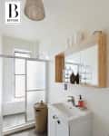 Small bathroom with a shower, wicker laundry basket, white sink, and wooden shelf with glass jars and decor.