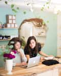 Two women working on a laptop at a wooden counter in a boutique with plants and shelves of products.