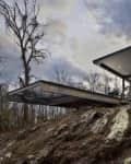 Modern glass and concrete house on rocky hillside surrounded by bare trees under a cloudy sky.