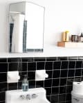 Bathroom with black tile backsplash, mirror, towel, shelf with toiletries, and toilet with paper rolls.