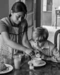 Woman helping a boy with breakfast at a wooden table, featuring pastries, milk, and a vintage radio in the background.