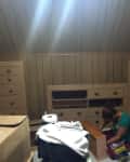 Attic room with wood paneling, woman assembling furniture, cardboard boxes, and scattered items.