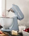 Blue stand mixer with whisk attachment on kitchen counter, surrounded by macarons, strawberries, butter, and measuring cups.
