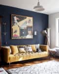 Living room with a mustard yellow sofa, eclectic artwork, wooden armoire, and a large window with plants.