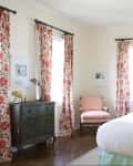 Bedroom with floral curtains, white bedding, dark dresser, and patterned chair.