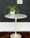 Marble-topped tulip table with a potted plant against a dark wall, small framed art above, and patterned rug below.