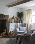 Cozy living room with a wood-burning stove, armchair, and plants, featuring a rustic mantel and light curtains.