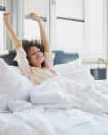 Person stretching in bed with white sheets, smiling, in a bright room with large windows and a black lamp.