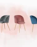 Four velvet dining chairs in gray, pink, blue, and red with wooden legs on a soft pink background.