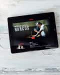 Tablet displaying Netflix series "Narcos" next to a cup of coffee and white earphones on a wooden table.